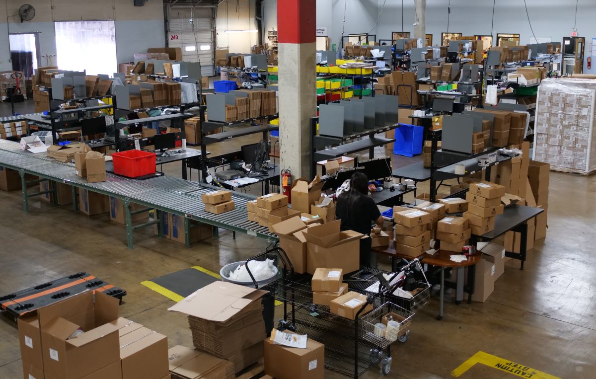 A large warehouse with boxes going down conveyor belts while a woman packs orders at her packing station.
