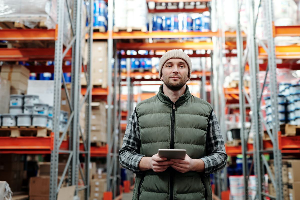 A man holding a tablet while standing in the aisle of a warehouse surrounded by goods.