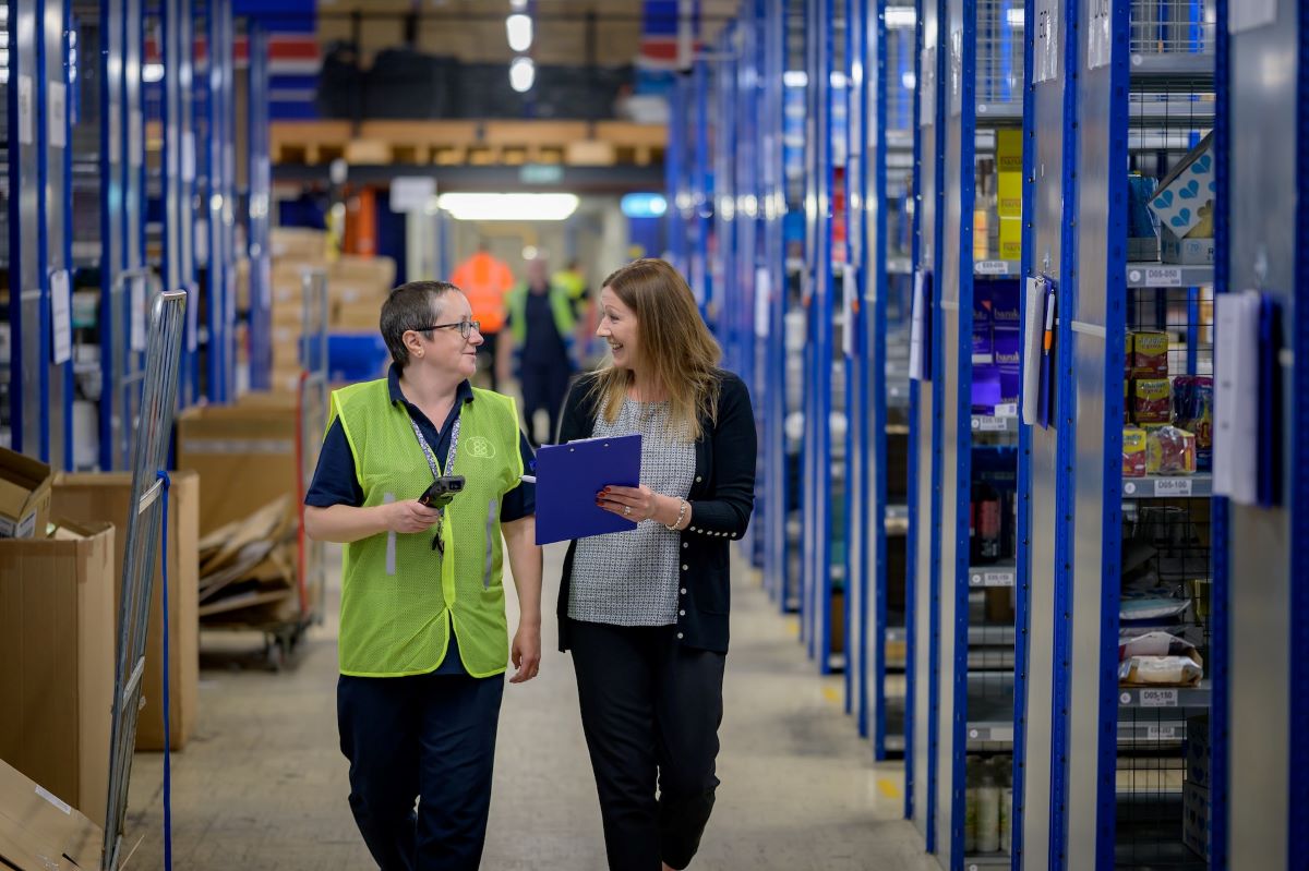 Two people walking down a warehouse aisle looking at a handheld device to count physical inventory.